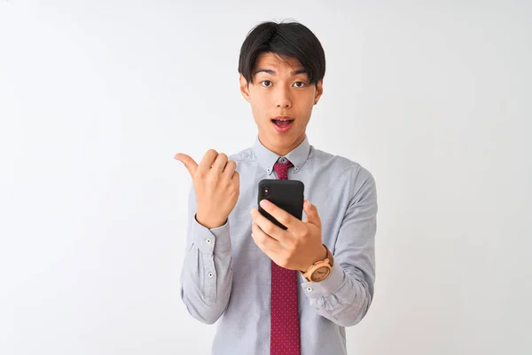 中国商人打着领带 用智能手机站在孤立的白色背景上 用大拇指指着旁边 笑容满面 — 图库照片