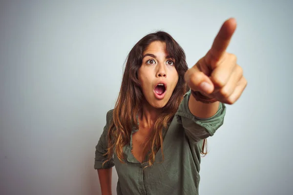 Young beautiful woman wearing green shirt standing over grey isolated background Pointing with finger surprised ahead, open mouth amazed expression, something on the front