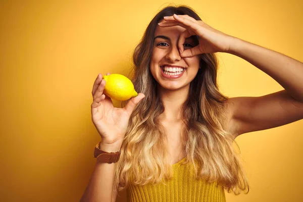 Young beautiful woman eating a lemon over yellow isolated background with happy face smiling doing ok sign with hand on eye looking through fingers