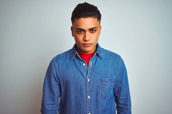 Young brazilian man wearing denim shirt standing over isolated white background with serious expression on face. Simple and natural looking at the camera.