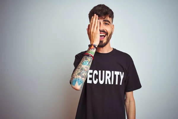 Young safeguard man with tattoo wering security uniform over isolated white background covering one eye with hand, confident smile on face and surprise emotion.