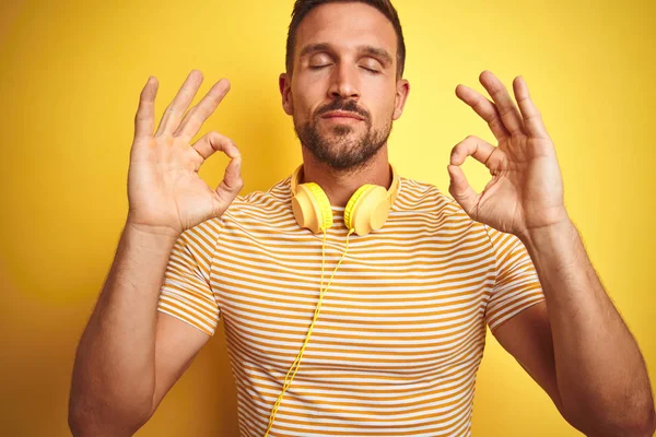 黄色の隔離された背景の上の音楽を聞くヘッドフォンを身に着けている若いハンサムな男は指で瞑想のジェスチャーをして閉じ目でリラックスし ヨガのコンセプト — ストック写真