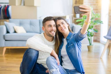 Yeni evin zemininde oturup akıllı telefon kullanarak selfie fotoğrafı çeken genç çift, yeni daireye taşındığı için mutlu bir şekilde gülümsüyor