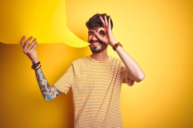 Dövmeli genç adam konuşma balonu tutuyor. Sarı arka planda duruyor. Gülümsüyor, gülümsüyor.