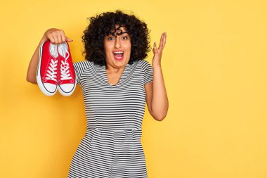 Kıvırcık saçlı genç Arap kadın izole edilmiş sarı arka planda spor ayakkabılarını tutuyor. Çok mutlu ve heyecanlı. Kazanan ifade zaferi büyük bir gülümsemeyle kutluyor ve ellerini kaldırıyor.