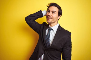 Takım elbiseli ve kravatlı genç yakışıklı adam izole edilmiş sarı arka planda duruyor. Gülümsüyor, saçlarına dokunuyor, el hareketi yapıyor, çekici ve modaya uygun poz veriyor.
