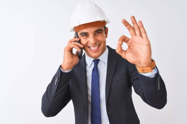 Miğfer takan genç mimar, izole edilmiş beyaz arka planda akıllı telefondan konuşuyor. Tamam işareti yapıyor, mükemmel bir sembol.