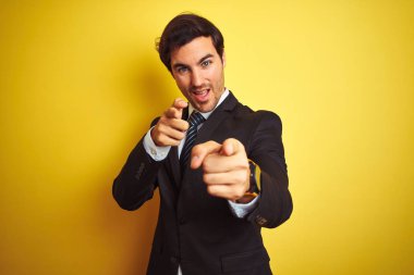 Takım elbiseli ve kravatlı genç yakışıklı işadamı izole edilmiş sarı arka planda durmuş mutlu ve komik yüzlü parmaklarını kameraya doğrultuyor. İyi enerji ve titreşimler.