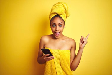 Duş üstüne havlu giyen Afro kadın izole edilmiş sarı arka plan üzerinde smatrphone kullanıyor. Çok mutlu bir şekilde elini ve parmağını kenarı işaret ediyor.