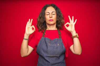 Kırmızı izole edilmiş arka planda önlük giyen orta yaşlı bir kadın rahatlamış ve parmaklarıyla meditasyon hareketi yaparken gözleri kapalı gülümsüyor. Yoga konsepti.