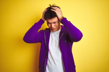 Mor kazak giyen Asyalı genç adam izole edilmiş sarı arka planda duruyor. Ağrı ve migren yüzünden baş ağrısı ve stresten muzdarip. Ellerini başının üstüne koy..