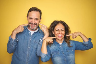 Güzel bir orta yaşlı çift, izole edilmiş sarı arka planda durup neşeli bir şekilde gülümseyerek parmakları ve ağızlarıyla işaret ediyorlar. Diş sağlığı konsepti.