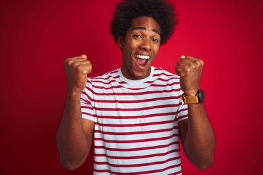 Afro saçlı, izole edilmiş kırmızı arka plan üzerine çizgili tişört giyen genç Afro Amerikalı adam kollarını kaldırarak ve açık gözlerle başarıyı kutluyor ve şaşırıyor. Kazanan konsept.