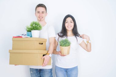 Young beautiful couple holding cardboard boxes standing over isolated white background with surprise face pointing finger to himself