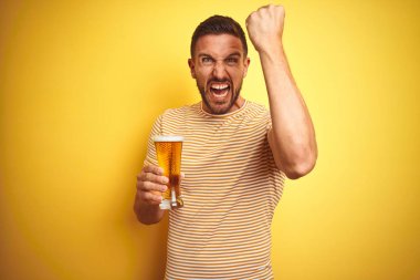 Genç yakışıklı adam izole edilmiş sarı arka planda bir bardak bira içerken sinirlenmiş ve sinirlenmiş öfkeyle bağırıyor, çıldırmış ve el kaldırarak bağırıyor, öfke kavramı