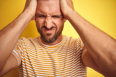Sarı izole edilmiş arka plan üzerine gündelik tişört giyen, acı ve migren yüzünden çaresiz ve stresli genç ve yakışıklı bir adam. Ellerini başının üstüne koy..