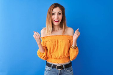 Beautiful redhead woman wearing orange casual t-shirt standing over isolated blue background celebrating surprised and amazed for success with arms raised and open eyes. Winner concept.