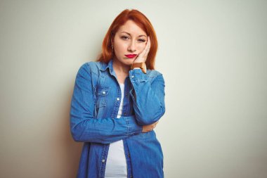 Genç, güzel, kızıl saçlı, kot gömlek giyen, izole edilmiş beyaz arka planda duran, yorgun ve bunalımda kollarını kavuşturmaktan sıkılmış görünen bir kadın..