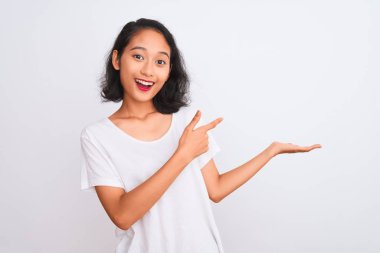 İzole edilmiş beyaz arka plan üzerinde duran gündelik tişört giyen genç Çinli kadın kameraya hayret ediyor ve gülümsüyor..