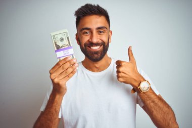 Young indian man holding dollars standing over isolated white background happy with big smile doing ok sign, thumb up with fingers, excellent sign