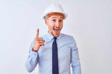 Young business man wearing contractor safety helmet over isolated background pointing finger up with successful idea. Exited and happy. Number one.