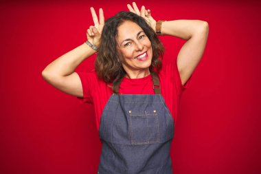 Kırmızı izole arka planda önlük giyen orta yaşlı bir kadın komik ve çılgın pozlar veriyor parmakları kafasında tavşan kulağı, neşeli gülüyor.