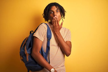 Afrika kökenli Amerikalı öğrenci sırt çantasıyla izole edilmiş sarı arka plan örtüsü ile ağzını kapatıyor. Yanlışlıkla utançla dolu ağzı, korkunun ifadesi, sessizlikten korkmak, gizli konsept.