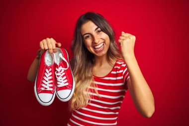 Young beautiful woman holding sneakers over red isolated background screaming proud and celebrating victory and success very excited, cheering emotion