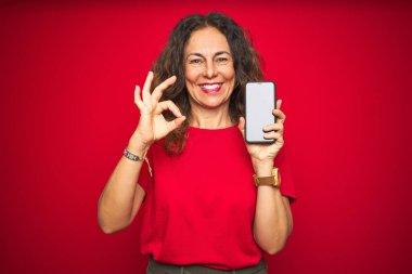 Kırmızı izole edilmiş arka planda akıllı telefon ekranı gösteren orta yaşlı bir kadın tamam işareti yapıyor, mükemmel bir sembol.