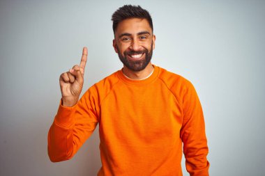 Young indian man wearing orange sweater over isolated white background showing and pointing up with finger number one while smiling confident and happy.
