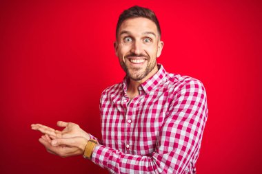 Kırmızı izole edilmiş arka plan üzerinde genç yakışıklı bir adam elleri açık, fotokopi alanını gösteren avuç içleri açık, gülümseyerek mutlu bir gülümseme sunuyor.