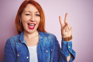 İzole edilmiş pembe arka plan üzerinde dikilen güzel, kızıl saçlı kadın kameraya bakıp gülümsüyor ve zafer işareti yapıyor. İki numara..