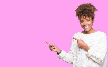 Gülümseyen ve iki elleri ve parmakları ile tarafına işaret kamera bakarak izole arka plan üzerinde kış kazak giyen güzel genç Afro-Amerikan kadın.