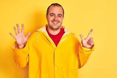 İzole edilmiş sarı arka planda duran yağmurluk giyen genç adam yedinci parmaklarını gösterip gülümserken kendinden emin ve mutlu görünüyor..