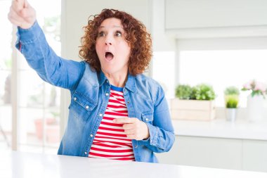 Kıvırcık saçlı, evde kot ceket giyen orta yaşlı bir kadın. Parmakları önde, ağzı açık, şaşırmış, ön tarafta bir şey var.