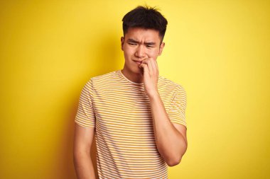 İzole edilmiş sarı arka planda duran tişört giyen Asyalı genç adam elleri ağız yiyen tırnaklarla gergin ve stresli görünüyor. Endişe sorunu.
