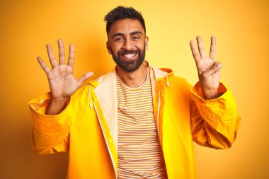 İzole edilmiş sarı arka planda duran yağmurluk giyen genç Hintli adam sekiz numaralı parmaklarıyla gülümseyerek kendinden emin ve mutlu bir şekilde işaret ediyor..