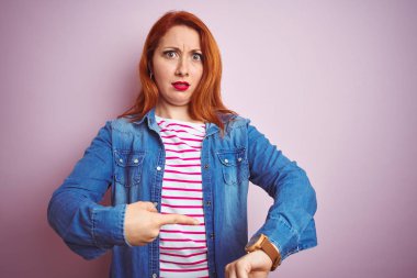 İzole edilmiş pembe arka plan üzerinde kot gömlek ve çizgili tişört giyen güzel kızıl saçlı kadın. Aceleyle saati, sabırsızlığı, son teslim tarihini geciktirdiği için üzgün ve kızgın olduğunu gösteriyor.