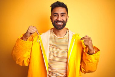 İzole edilmiş sarı arka planda duran yağmurluk giyen genç Hintli adam yüzünde bir gülümsemeyle gururlu ve mutlu bir şekilde kendini işaret ediyor..