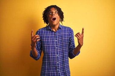 Rastgele gömlek giyen Afro adam sarı arka planda dikilmiş şaşkın şaşkın yukarı bakıyor, parmaklarıyla işaret ediyor ve kollarını kaldırıyordu..