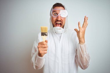 Profesyonel işçi teçhizatı giyen ressam arka planda fırça tutuyor çok mutlu ve heyecanlı, kazanan yüz ifadesi zaferi büyük bir gülümsemeyle kutluyor ve ellerini kaldırıyor.