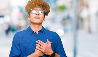 Afro saçlı mavi gözlük ile gülümseyen genç yakışıklı adam kapalı gözler ve yüzünde minnettar jest ile göğüs üzerinde eller. Sağlık kavramı.