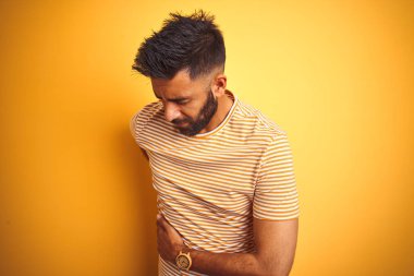 İzole edilmiş sarı arka planda duran tişörtlü genç Hintli adam sırt ağrısı çekiyor, elleriyle dokunuyor, kas ağrısı çekiyor.