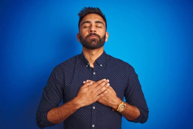 İzole edilmiş mavi arka plan üzerinde duran gömlek giyen genç Hintli zarif adam elleri göğsünde, gözleri kapalı ve yüzünde minnet dolu bir jest ile gülümsüyor. Sağlık kavramı.