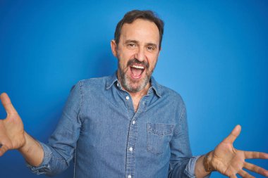 Yakışıklı, orta yaşlı, gri saçlı, soyutlanmış mavi arka planda neşeli gülümseyen, dost canlısı, olumlu ve kendinden emin bir selamlama.