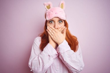 Pembe izole arka planda pijama giyen güzel, kızıl saçlı bir kadın yanlışlıkla ağzı ellerle kapatmış. Gizli konsept.