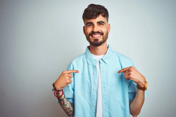 Young man with tattoo wearing blue shirt standing over isolated white background looking confident with smile on face, pointing oneself with fingers proud and happy.