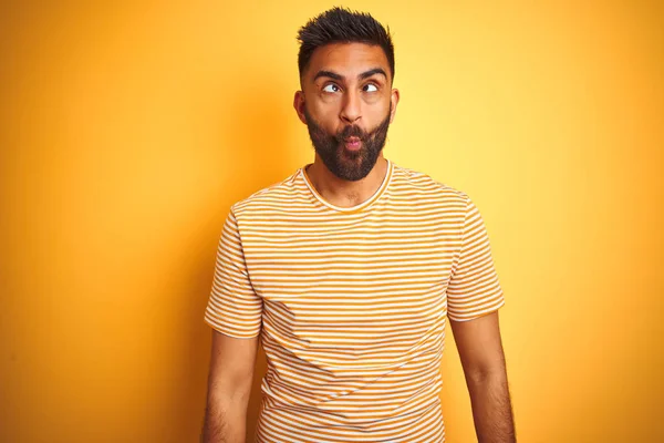 Young indian man wearing t-shirt standing over isolated yellow background making fish face with lips, crazy and comical gesture. Funny expression.