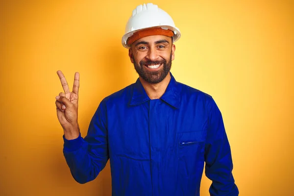 勝利の印をしているカメラで幸せな顔のウィンクで微笑む孤立した黄色の背景の上に制服とヘルメットを身に着けているハンサムなインドの労働者の男 第二番 — ストック写真