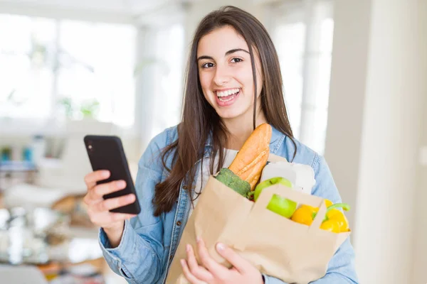 Young woman holding a paper bag full of fresh groceries and using smartphone app for supermarket delivery